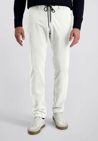 Witte corduroy broek met een gestructureerde afwerking, slim fit, en zwarte trekkoord in de taille. Gecombineerd met lichte sneakers met beige accenten.