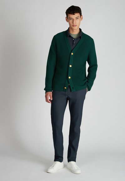 Zielony sweter z dzianiny z dekoltem w serek i złotymi guzikami, w połączeniu z granatowymi spodniami i białymi trampkami. Gładka tekstura i luźny krój.