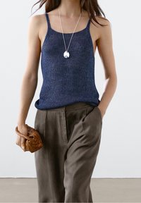 Donkerblauw gebreid tanktop met spaghettibandjes; gecombineerd met hoog-waist olijfgroene broek. Het model houdt een kleine geweven tas vast.