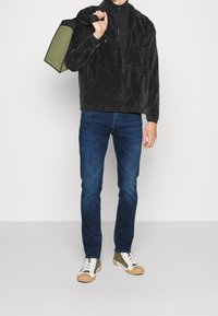 Svart vaxad jacka med framfickor, svart zip-up topp, blå jeans och gröna & vita sneakers med strukturerade gummisulor.