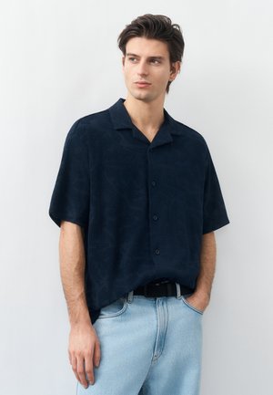 Νεαρός άνδρας που φορά σκούρο μπλε κοντομάνικο πουκάμισο με κουμπιά και ανοιχτόχρωμο τζιν, στέκεται με το ένα χέρι στην τσέπη μπροστά σε λευκό φόντο.