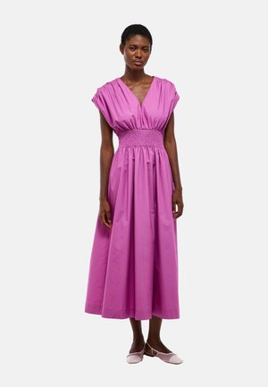 Femme debout, portant une robe midi sans manches rose vif avec une taille froncée et un décolleté en V, assortie à des chaussures plates roses.