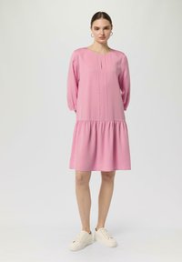 Rosa, knielanges, langärmliges Kleid mit einem gerafften Rock, ausgestattet mit einem runden Ausschnitt und kleinen Fältchen im Brustbereich. Kombiniert mit hellen Sneakers.