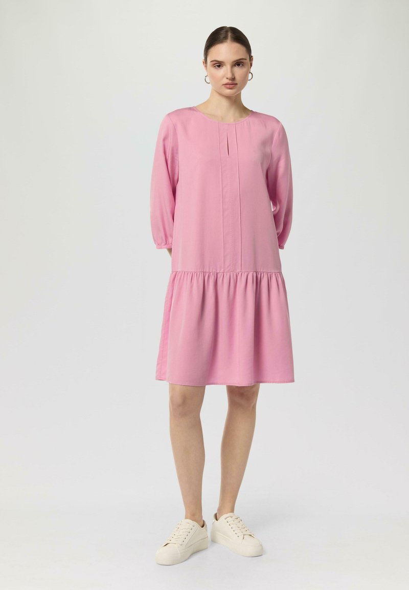 Rosa, knielanges, langärmliges Kleid mit einem gerafften Rock, ausgestattet mit einem runden Ausschnitt und kleinen Fältchen im Brustbereich. Kombiniert mit hellen Sneakers.