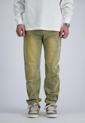 Man wearing light green faded jeans, white long-sleeve shirt, white and beige sneakers, and a silver wristwatch.