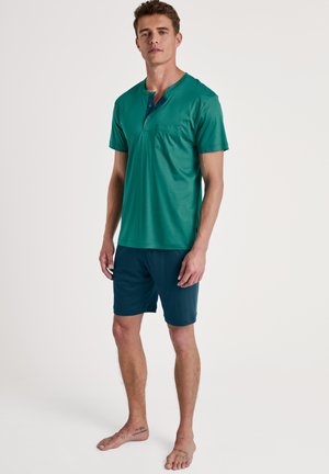Groene kortebloes met een zak en een drie-knopen sluiting, gecombineerd met donkerblauwe shorts van zacht, lichtgewicht stof.