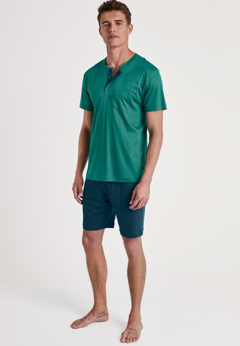 Groene kortebloes met een zak en een drie-knopen sluiting, gecombineerd met donkerblauwe shorts van zacht, lichtgewicht stof.