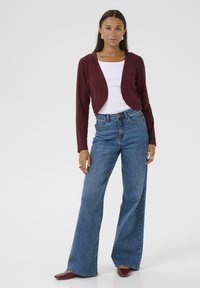Cardigan court bordeaux porté sur un top blanc, associé à un jean large bleu clair taille haute et des chaussures bordeaux. Modèle debout.