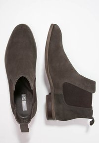 Botas Chelsea de ante marrón oscuro con paneles elásticos en los laterales, tiradores en los talones y tacones de madera bajos y apilados, mostradas desde vistas superior y lateral.