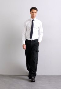 Camicia bianca a maniche lunghe infilata dentro, pantaloni cargo neri con tasche e scarpe nere. Design semplice e sartoriale in colori neutri.