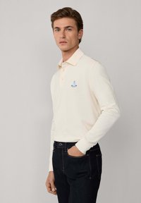 Polo a maniche lunghe color crema con colletto, dotato di un logo ricamato blu. Abbinato a jeans scuri, evidenziando il design casual.