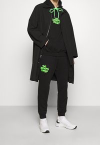 Person som bär en svart huvtröja med neongrön text "The Iceberg Family", svart jacka och vita sneakers mot en enhetlig bakgrund.