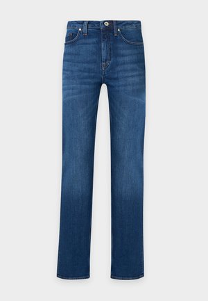 Un mannequin se tient devant un fond uni, vêtu d’un col roulé blanc, d’un cardigan bleu marine, d’un jean bleu, de bottines noires et d’un sac porté à l’épaule.