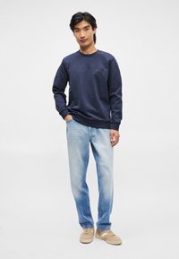 Sweatshirt azul-marinho com punhos e bainha canelados, apresentando um padrão texturizado subtil e um pequeno logo no peito. Calças jeans azul-claro e ténis castanhos.