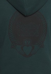 Mörkgrön tyg med en svart emblem som visar två händer som håller ett hjärta under texten "See By Chloé" inuti en cirkulär flätad kant.