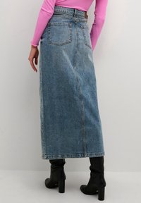 Falda maxi de mezclilla en azul claro, con cintura alta, dos bolsillos traseros y corte recto. Se lleva con botas negras de tacón alto.