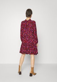 Sukienka z czarnego materiału z żywym wzorem kwiatowym w odcieniach różu i czerwieni, z długimi rękawami, falbaną u dołu i szczegółem z wiązaniem przy szyi. Noszona z beżowymi botkami do kostek.