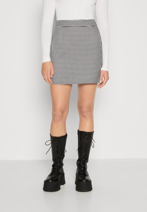 Frau trägt einen schwarz-weiß karierten Minirock im Hahnentrittmuster, ein weißes langärmeliges Oberteil und kniehohe schwarze Schnürstiefel mit Plateau.