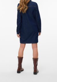 Femme debout de dos portant une robe en jean bleu foncé à manches longues et des bottes en cuir marron mi-mollet sur fond blanc.