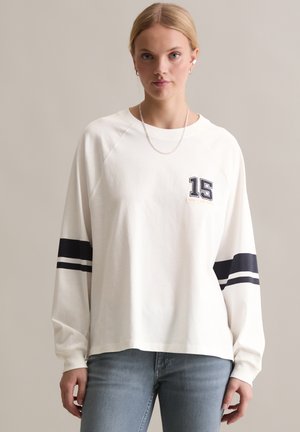 White sweatshirt with long raglan sleeves, navy stripes on the sleeves, and a "15" graphic on the chest. Casual fit and soft texture.