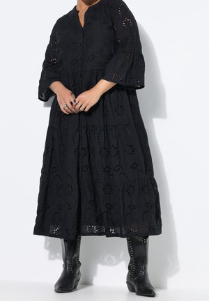 Personne portant une robe midi noire brodée avec des manches trois-quarts et des bottes noires cloutées mi-mollet, debout sur un fond blanc.