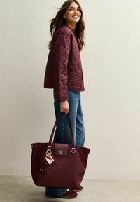 Mörkröd tote-väska med läderdetaljer, dragkedjeslutning och guldfärgad metalldetaljer, prydd med en dekorativ nyckelring. Bärs med en quiltad mörkröd jacka och jeans.