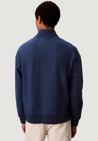 Chaqueta tipo pulóver azul marino con cuello, puños y cintura acanalados. Tejido suave y texturizado con un ajuste relajado, sin costuras visibles.