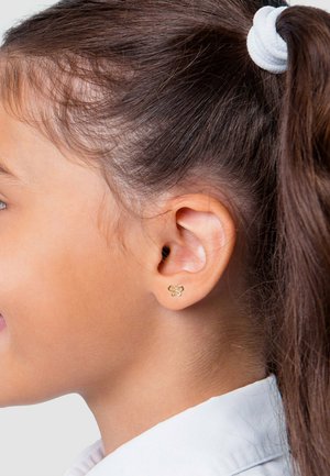 Junges Mädchen mit dunklem Haar, das nach hinten gebunden ist. Sie trägt kleine goldene Ohrringe in Schmetterlingsform und ein weißes Hemd mit Kragen.