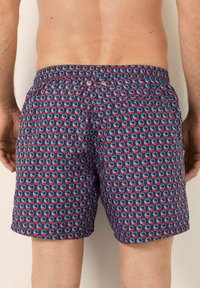 Mies, der marineblaue Badehosen mit rotem und hellblauem geometrischem Muster trägt, von hinten vor einer schlichten Wand gezeigt.