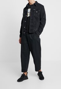 Černá džínová bunda s dvěma kapsami na hrudi, černé tričko s potiskem, volné černé kalhoty a černé tenisky, vše v jednotném černobílém stylu.