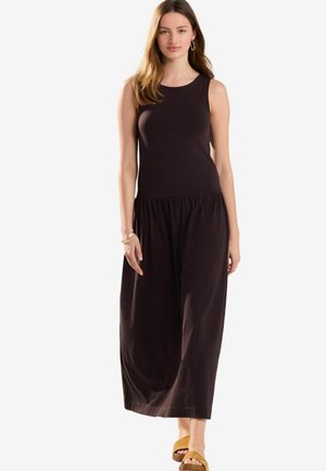 Robe sans manches marron foncé avec un haut ajusté et une taille froncée, tombant jusqu'aux chevilles. Modèle portant des sandales marron clair.