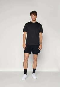 Tricou negru cu mânecă scurtă, combinat cu pantaloni scurți negri. Întreaga ținută are o croială lejeră și este completată de adidași albi și șosete închise la culoare.