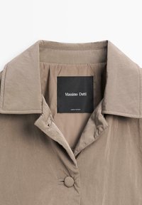 Lichtbruine jacket met een soepele textuur, voorzien van een klassieke kraag, knoopsluiting en een label met de tekst "Massimo Dutti" aan de binnenkant.