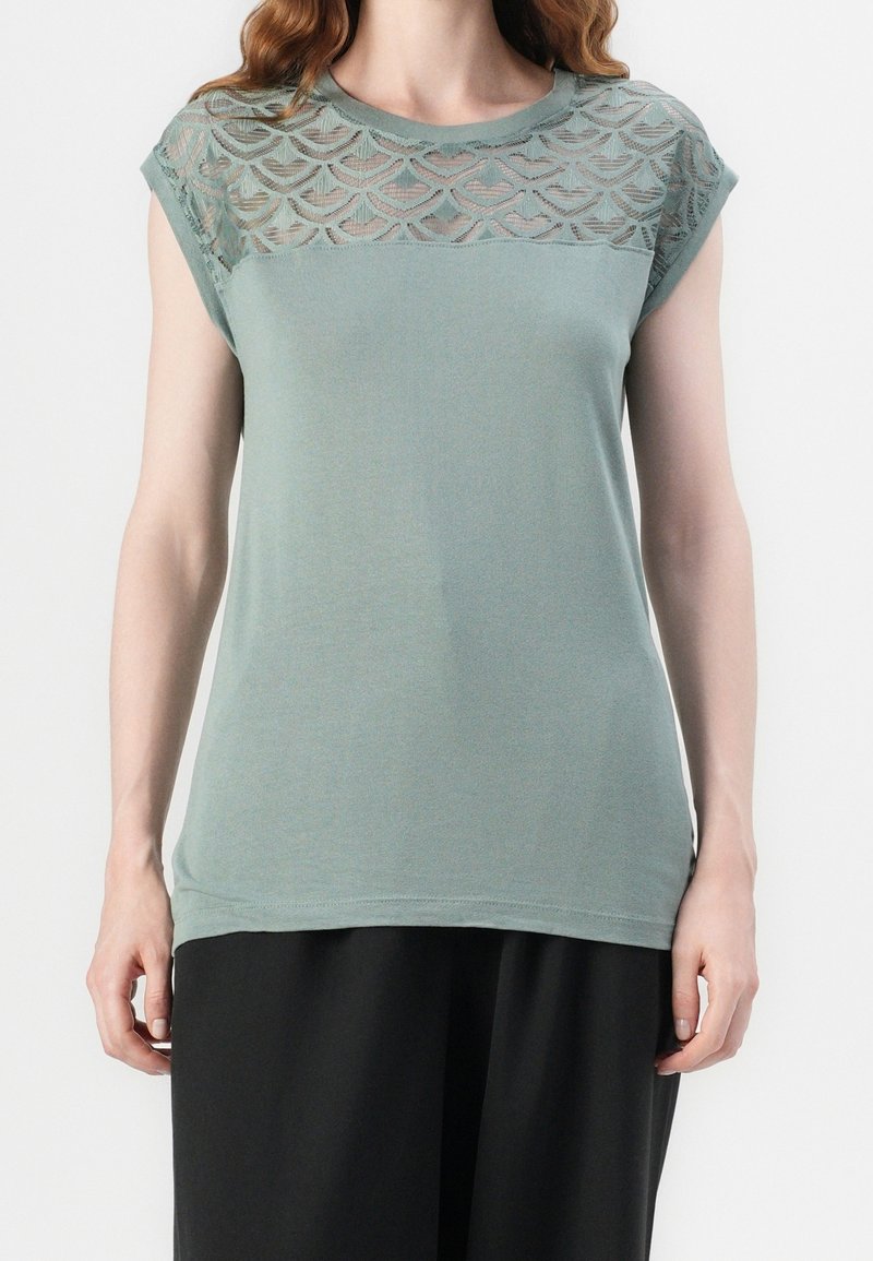 Femme portant un haut vert sauge sans manches avec un empiècement en dentelle présentant un motif en coquilles, associée à un pantalon noir.
