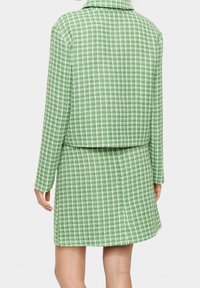 Zelený sada saka a sukně z tvídu s kostkovaným vzorem, strukturovaným límcem a dlouhými rukávy. Texturovaná látka s bílými akcenty.