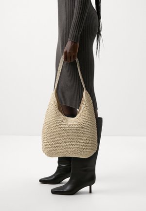 Beige Schultertasche aus gewebtem Stroh mit weicher, strukturierter Oberfläche und breitem Riemen, gehalten von einer Person in einem enganliegenden grauen gerippten Kleid und schwarzen Stiefeln.