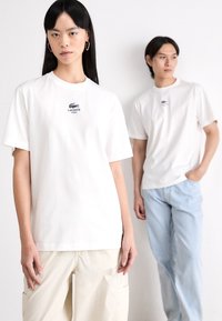 Hvite bomull crew neck t-skjorter med brodert blå Lacoste-logo. En modell i lyse beige bukser; den andre i lyse blå jeans.