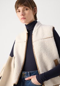 Beige fleece vest with a high collar, zip closure, and brown accents; worn over a navy turtleneck, paired with blue denim jeans.