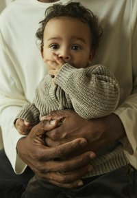 Pull en tricot beige pour bébé avec des poignets côtelés, tenu par une personne portant une chemise crème à manches longues. Des mains foncées contrastent avec le tissu clair.