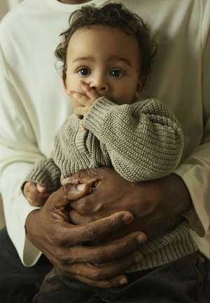 Beige Strickpullover für Babys mit gerippten Bündchen, gehalten von einer Person in einem cremefarbenen Langarmshirt. Dunkle Hände heben sich kontrastreich vom hellen Stoff ab.