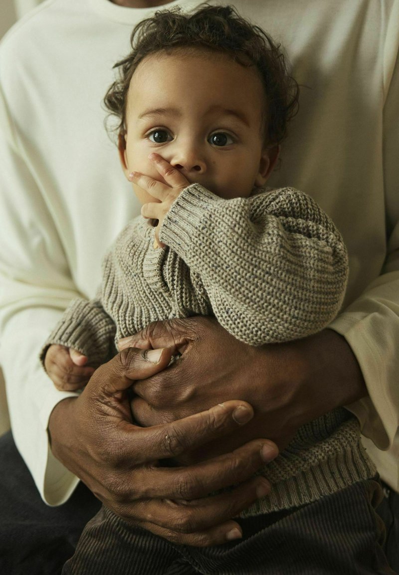 Pull en tricot beige pour bébé avec des poignets côtelés, tenu par une personne portant une chemise crème à manches longues. Des mains foncées contrastent avec le tissu clair.