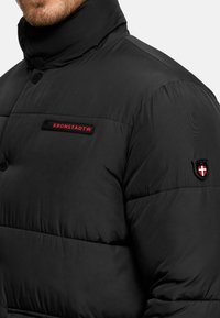 Man draagt een zwarte donzen jas met hoge kraag, rode "KRONSTADT" logo op de borst en schildembleem op de bovenste mouw.