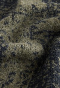 Teksturirana volna v olivno zeleni in črni barvi, z madežastim vzorcem, vidnimi vlakni ter mehkim, toplim videzom.