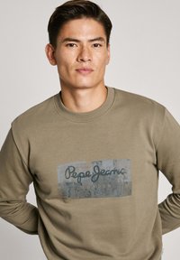 Sudadera verde oliva con cuello redondo, que presenta un logo rectangular gris impreso en la parte frontal, hecha de suave material de algodón.