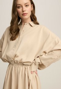 Beżowa oversize koszula z kołnierzykiem, zebranym w pasie i szerokimi rękawami, wykonana z miękkiego materiału o gładkiej fakturze.