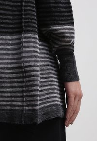 Randig tröja med en gradient från mörkgrå till ljusgrå, med en texturerad väv och en något oversized passform, där en hand vilar i närheten.