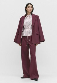 Maroon tailored blazer, loose-fit trousers, and a light striped blouse with a drawstring waist. Smooth fabric with structured silhouette.