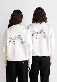 Sweatshirt crème avec un design éclaboussé coloré et "MILANO" en lettres majuscules dans le dos. Assorti avec un pantalon noir.