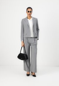 Grå skreddersydd dress med en kort jakke, vide bukser og en hvit skjorte. Svart håndveske med en buet form. Cat-eye solbriller.