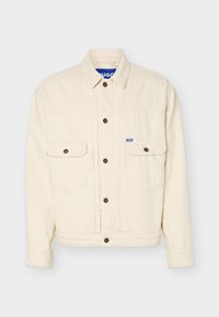 Μπεζ μακρυμάνικο μπουφάν με κουμπωτή μπροστινή πλευρά, δύο τσέπες στο στήθος με καπάκι, γιακά και μικρή μπλε ετικέτα με λογότυπο στη δεξιά τσέπη.